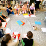 Na zdjęciu dzieci siedzące w kółku na ziemi przed nimi prowadzący . Na ziemi przed kazdym dzieckiem kolorowe kartki A4. Za nimi regały a na nich ksiazki.