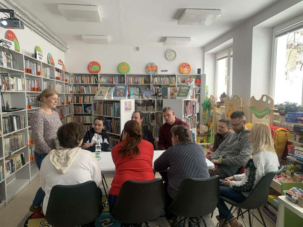 na zdjęciu sala biblioteczna a w niej stół przy którym siedza mężczyźni i kobiety uczestniczące w warsztatach kreatywno - literackich. Wokół rekały z książkami.