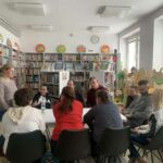 na zdjęciu sala biblioteczna a w niej stół przy którym siedza mężczyźni i kobiety uczestniczące w warsztatach kreatywno - literackich. Wokół rekały z książkami.