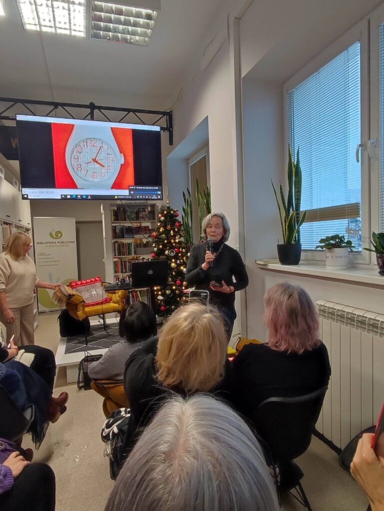 Na zdjęciu wnętrze biblioteki w którym odbywa się spotkanie autorskie z Ewą Chotomską. Na krzesłach tyłem siedza osoby, które przybyły na spotkanie. Z przodu widać kobietę , która przemawia.