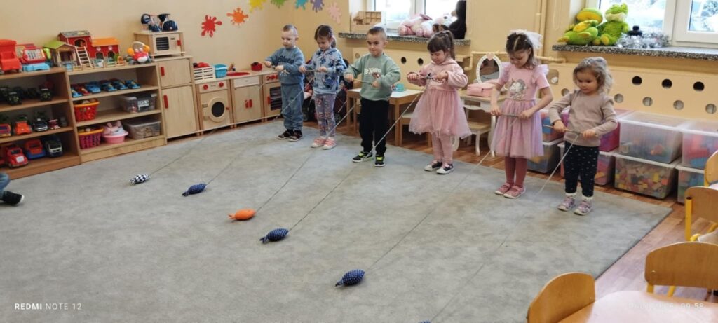 Szóstka przedszkolaków stoi w rzędzie. W rękach trzymają patyczki na których są sznurki i na końcu sznurka jest rybka. Dzieci bawią sie w łowienie rybek.