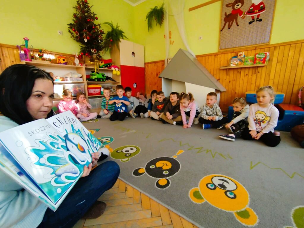 Na zdjęciu [ani bibliotekarka trzymająca otwartą książkę. Wokoł niej na ziemi siedzą dzieci w sali w przedszkolu na kolorowym dywanie.
