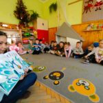 Na zdjęciu [ani bibliotekarka trzymająca otwartą książkę. Wokoł niej na ziemi siedzą dzieci w sali w przedszkolu na kolorowym dywanie.