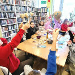 Przy dużym stole siedzi grupa dziesięciu dzieci i trzymaja w ręku wykonane przez siebie serca. Wokół regały z książkami.