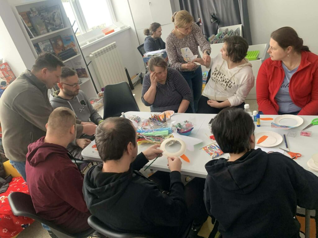 Przy stole siedzą osoby kobiety i mężczyźni, którzy przybyli na warsztaty kreatywno - literackie. Wspólnie wykonuja pracę plastyczną bałwankową maskę. Na stole kolorowe materiały potrzebne do warsztatów.