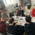 Przy stole siedzą osoby kobiety i mężczyźni, którzy przybyli na warsztaty kreatywno - literackie. Wspólnie wykonuja pracę plastyczną bałwankową maskę. Na stole kolorowe materiały potrzebne do warsztatów.