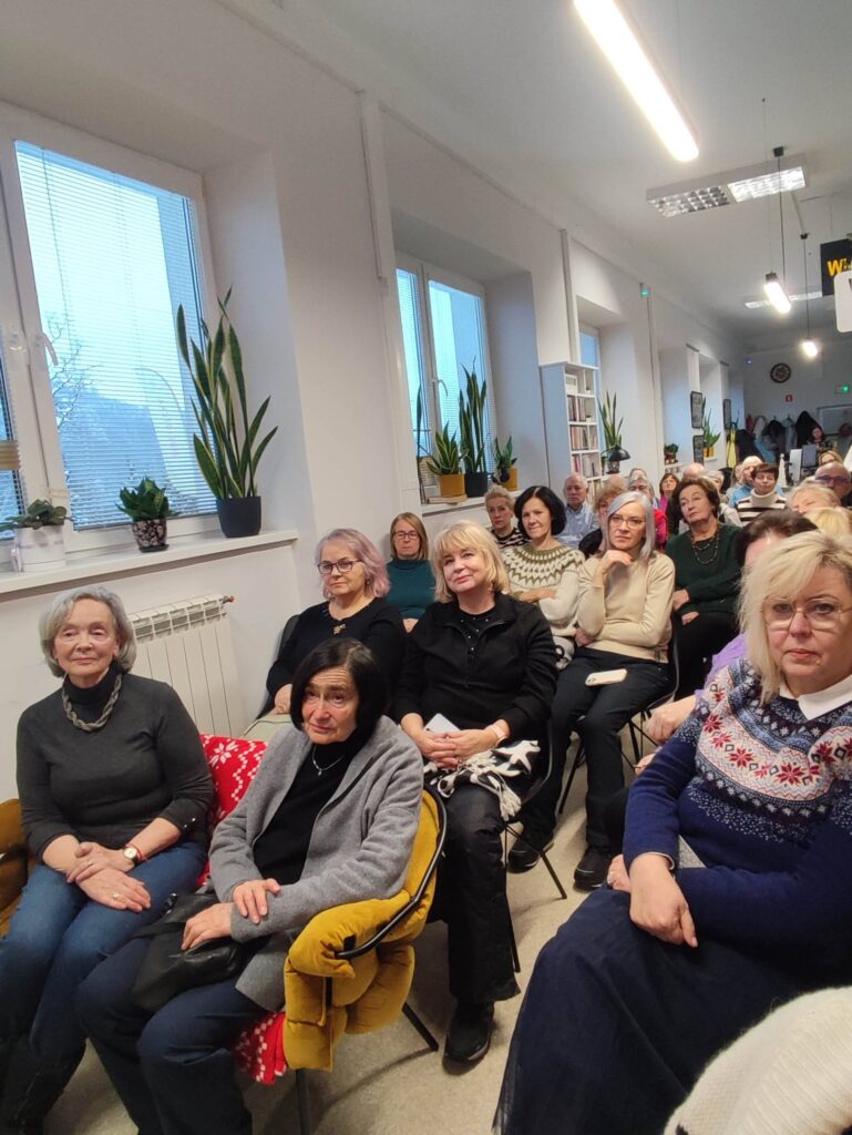 Na zdjęciu wnętrze biblioteki w którym odbywa się spotkanie autorskie z Ewą Chotomską. Na krzesłach przodem siedza osoby, które przybyły na spotkanie.