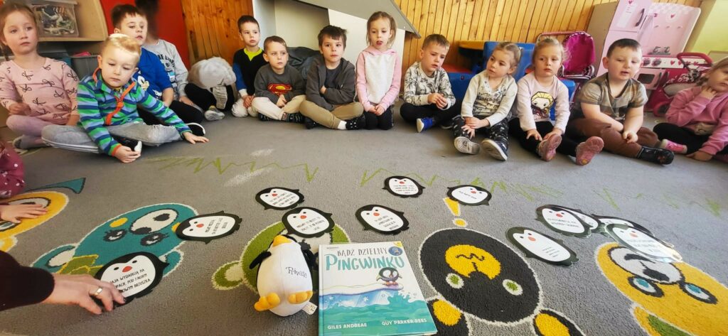 Na zdjęciu dzieci siedzące na kolorowym dywanie . Przed nimi książka o Pingwinku.