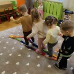 Czwórka dzieci przedszkolnych uczestniczy w zabawie. Przechodza po kolorowej taśmie. Z boku z prawej strony stoi pani w ręku z telefonem. a obok niej mała dziewczynka.