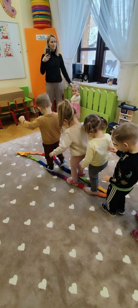 Czwórka dzieci przedszkolnych uczestniczy w zabawie. Przechodza po kolorowej taśmie. Z boku z prawej strony stoi pani w ręku z telefonem. a obok niej mała dziewczynka.