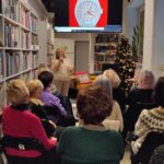 Na zdjęciu wnętrze biblioteki w którym odbywa się spotkanie autorskie z Ewą Chotomską. Na krzesłach tyłem siedza osoby, które przybyły na spotkanie. Z przodu widać kobietę , która przemawia.