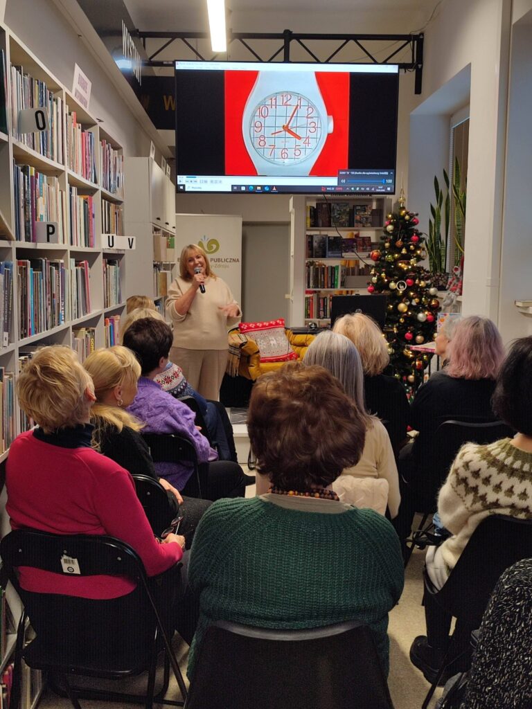 Na zdjęciu wnętrze biblioteki w którym odbywa się spotkanie autorskie z Ewą Chotomską. Na krzesłach tyłem siedza osoby, które przybyły na spotkanie. Z przodu widać kobietę , która przemawia.