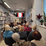 Na zdjęciu wnętrze biblioteki w którym odbywa się spotkanie autorskie z Ewą Chotomską. Na krzesłach tyłem siedza osoby, które przybyły na spotkanie. Z przodu widać kobietę , która przemawia.