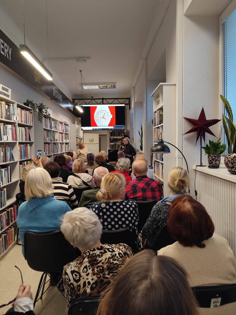 Na zdjęciu wnętrze biblioteki w którym odbywa się spotkanie autorskie z Ewą Chotomską. Na krzesłach tyłem siedza osoby, które przybyły na spotkanie. Z przodu widać kobietę , która przemawia.