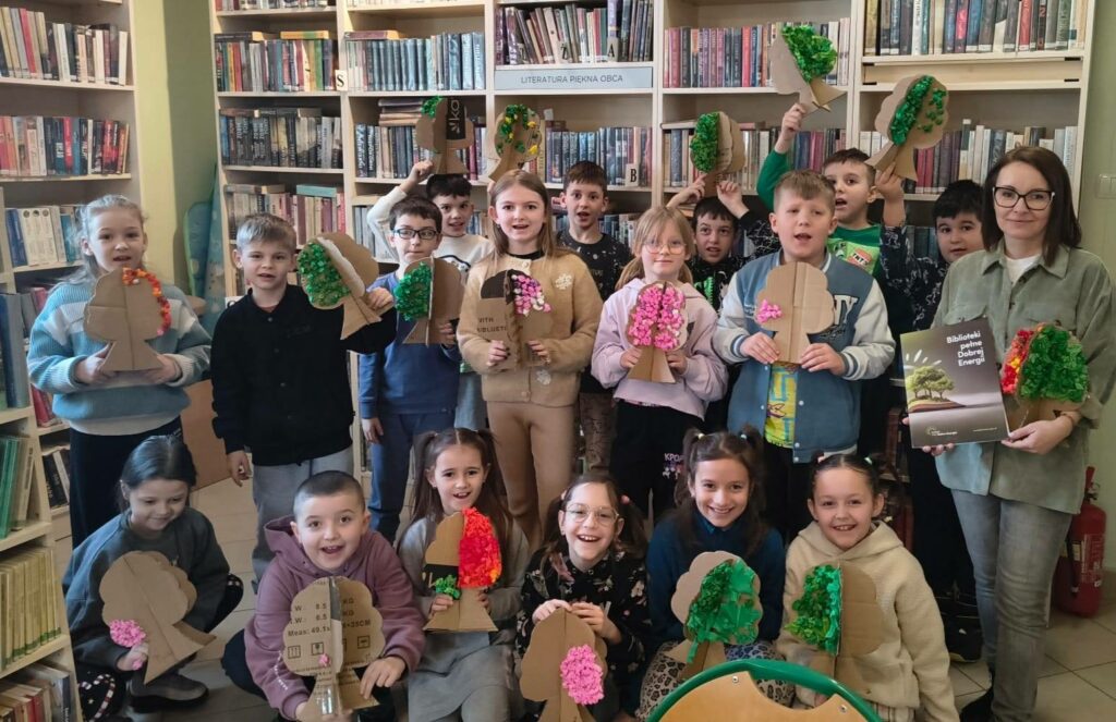 Dzieci wraz z panią biblotekarką stoja na tle regałuu i prezentuja wykonane przez siebie tekturowe drzewa.