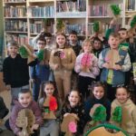 Dzieci wraz z panią biblotekarką stoja na tle regałuu i prezentuja wykonane przez siebie tekturowe drzewa.