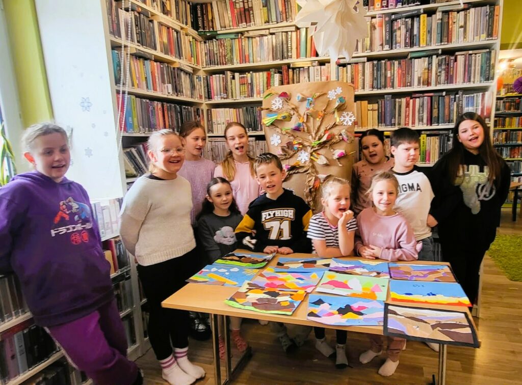 na zdjęciu widać dzieci stojące na tle regału z książkami a przednimi na stole wykonane przez nich prace plastyczne