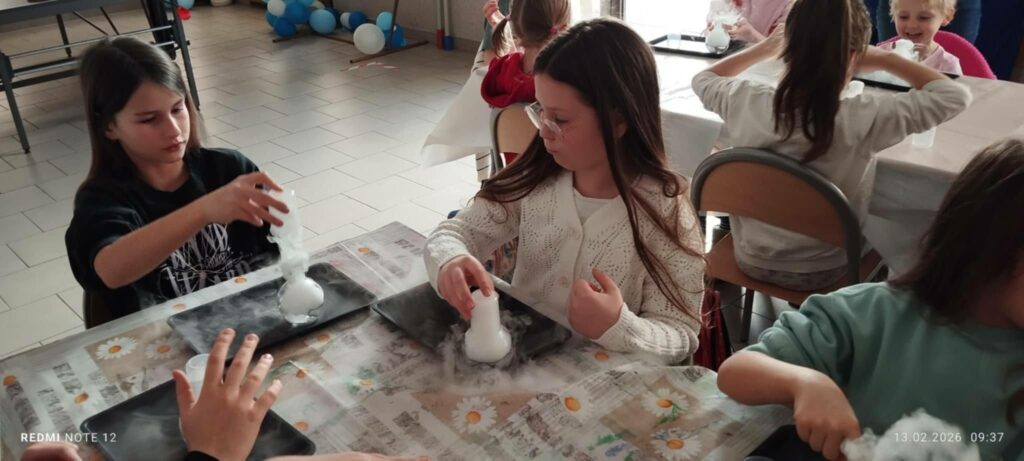 Przy stole siedzą dwie dziewczynki i wykonują eksperymenty.