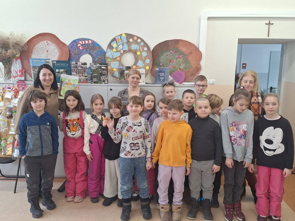 Do zdjęcia ustawiła się klasa szesnaście dzieci i trzy panie. Dzieci ubrane kolorowo. Jedna z pan trzym a ręku ksiązke i prezentuję ją