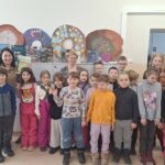 Do zdjęcia ustawiła się klasa szesnaście dzieci i trzy panie. Dzieci ubrane kolorowo. Jedna z pan trzym a ręku ksiązke i prezentuję ją