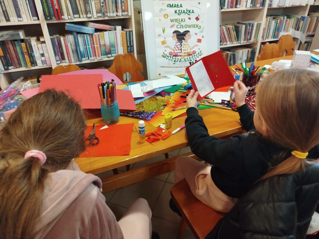Przy stle w sali bibliotecznej siedzą dwie dziewczynki i wykonuja prace plastyczne - kartki walentynkowe