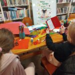 Przy stle w sali bibliotecznej siedzą dwie dziewczynki i wykonuja prace plastyczne - kartki walentynkowe