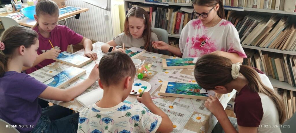 Dzieci siedzą przy stole i wykonują na drewnie pracę platyczna pejzaż