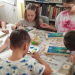 Dzieci siedzą przy stole i wykonują na drewnie pracę platyczna pejzaż