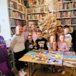 na zdjęciu widać dzieci stojące na tle regału z książkami a przednimi na stole wykonane przez nich prace plastyczne