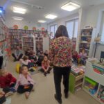Na zdjęciu kolorowa sala biblioteczna a w niej bibliotekarka , która prowadzi zajęćia feryjne z dziećmi. Dzieci siedza przed nią na podłodze. Wokół kolorowe regały z ksiązkami.