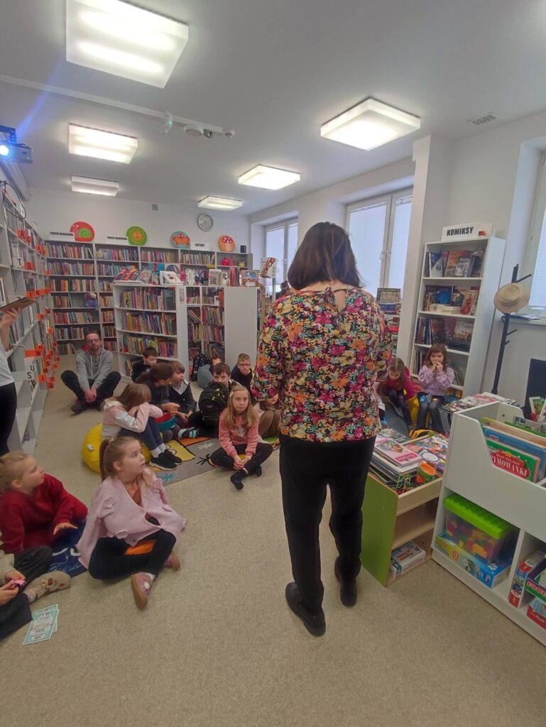 Na zdjęciu kolorowa sala biblioteczna a w niej bibliotekarka , która prowadzi zajęćia feryjne z dziećmi. Dzieci siedza przed nią na podłodze. Wokół kolorowe regały z ksiązkami.