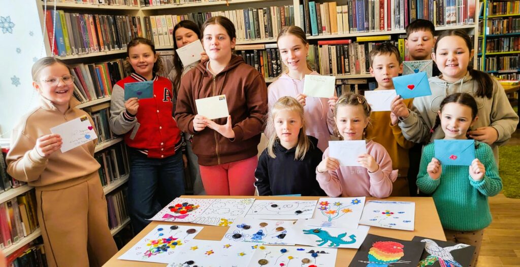 na zdjęciu widać dzieci w sali bibliotecznej. Stoja na tle ragałów i trzymaja w ręku zaadresowane koperty ze znaczkami, które wyślą pocztą. Przed nimi na stole kolorowe prace plastyczne