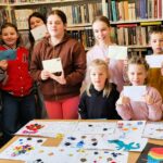 na zdjęciu widać dzieci w sali bibliotecznej. Stoja na tle ragałów i trzymaja w ręku zaadresowane koperty ze znaczkami, które wyślą pocztą. Przed nimi na stole kolorowe prace plastyczne