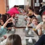 Przy dwóch długich stołach siedzą dzieci ze szkoły podstawowej i wykonują eksperymeny . Trzy panie pomagają dzieciom w eksperymencie.