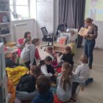 W sali bibliotecznej na ziemi i pufach siedza dzieci ze szkoły podstawowej a pani bibliotekarka czyta im ksiązkę.