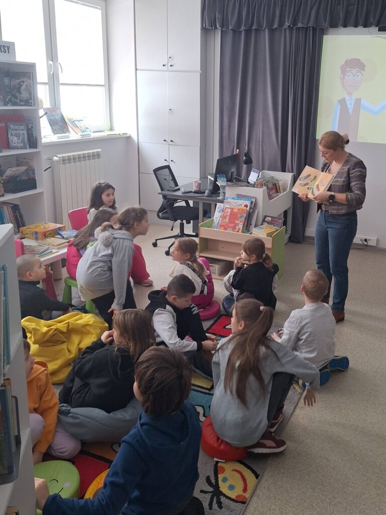 W sali bibliotecznej na ziemi i pufach siedza dzieci ze szkoły podstawowej a pani bibliotekarka czyta im ksiązkę.
