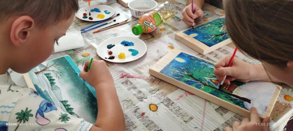 Dzieci siedzą przy stole i wykonują na drewnie pracę platyczna pejzaż