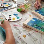 Dzieci siedzą przy stole i wykonują na drewnie pracę platyczna pejzaż