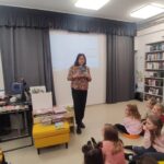 W sali bibliotecznej stoi bibliotekarka i prezentuje ksiązki. Przed nią dzieci siedzą na ziemi.