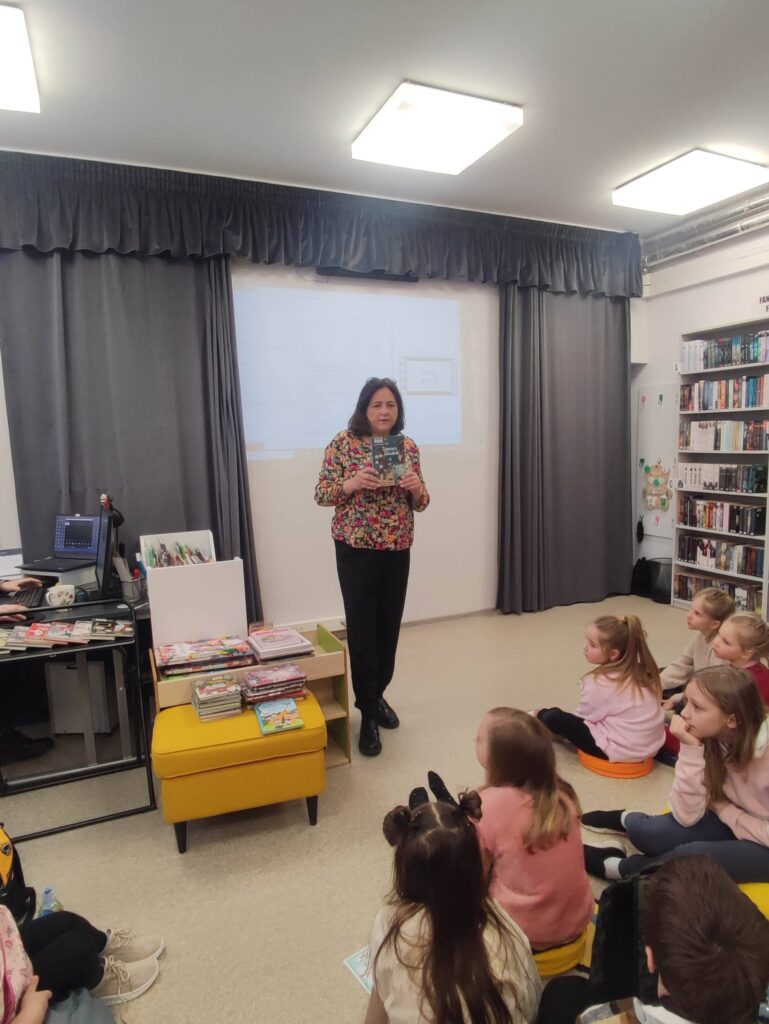 W sali bibliotecznej stoi bibliotekarka i prezentuje ksiązki. Przed nią dzieci siedzą na ziemi.