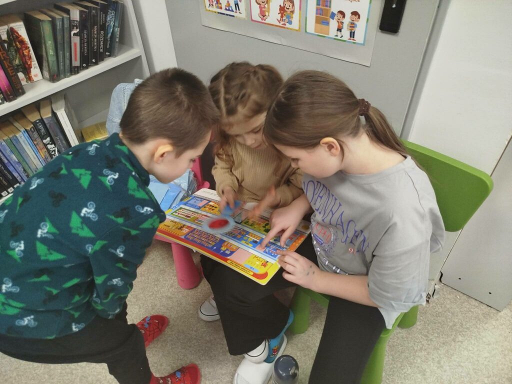 na krzesełkach siedzą dwie dziewczynki a przy nich pochylony stoi jeden chłopiec. W trójke oglądaja ksiązeczkę .