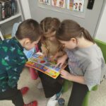 na krzesełkach siedzą dwie dziewczynki a przy nich pochylony stoi jeden chłopiec. W trójke oglądaja ksiązeczkę .