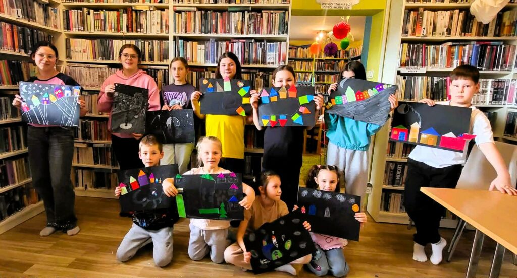 na zdjęciu widać prace plastyczne wykonane przez dzieci. Dzieci stoja na tle regału i trzymają w ręku prace plastyczne