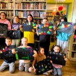 na zdjęciu widać prace plastyczne wykonane przez dzieci. Dzieci stoja na tle regału i trzymają w ręku prace plastyczne