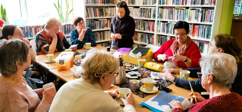 na zdjęciu widać panie seniorki siedzące wokół stołu w sali bibliotecznej i wykonujące korale