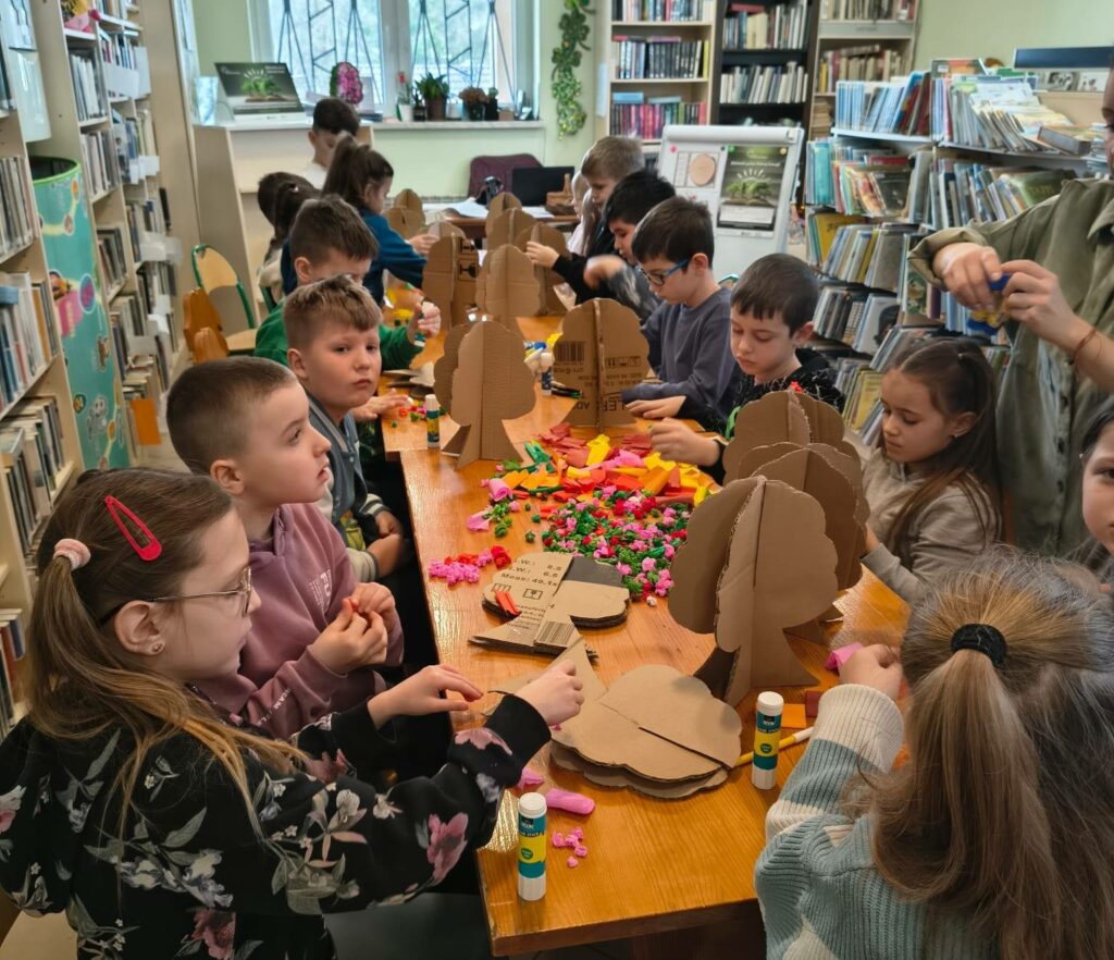 Przy długim stole w sali bibliotecznej siedza dzieci w w ieku szkoły podstawowej . Dzieci wykonuja drzewa. Tekturowe drzewa oklejaja bibułą.