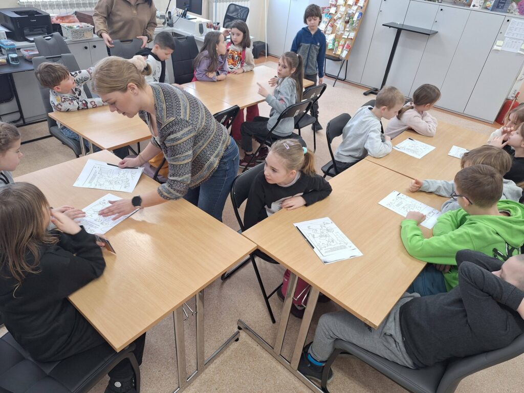 W sali biblioteczne przy stołach siedza dzoeci w wieku początkowej szkoły podstawowej. Przed niki kartki z zagadkami