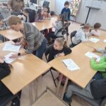 W sali biblioteczne przy stołach siedza dzoeci w wieku początkowej szkoły podstawowej. Przed niki kartki z zagadkami