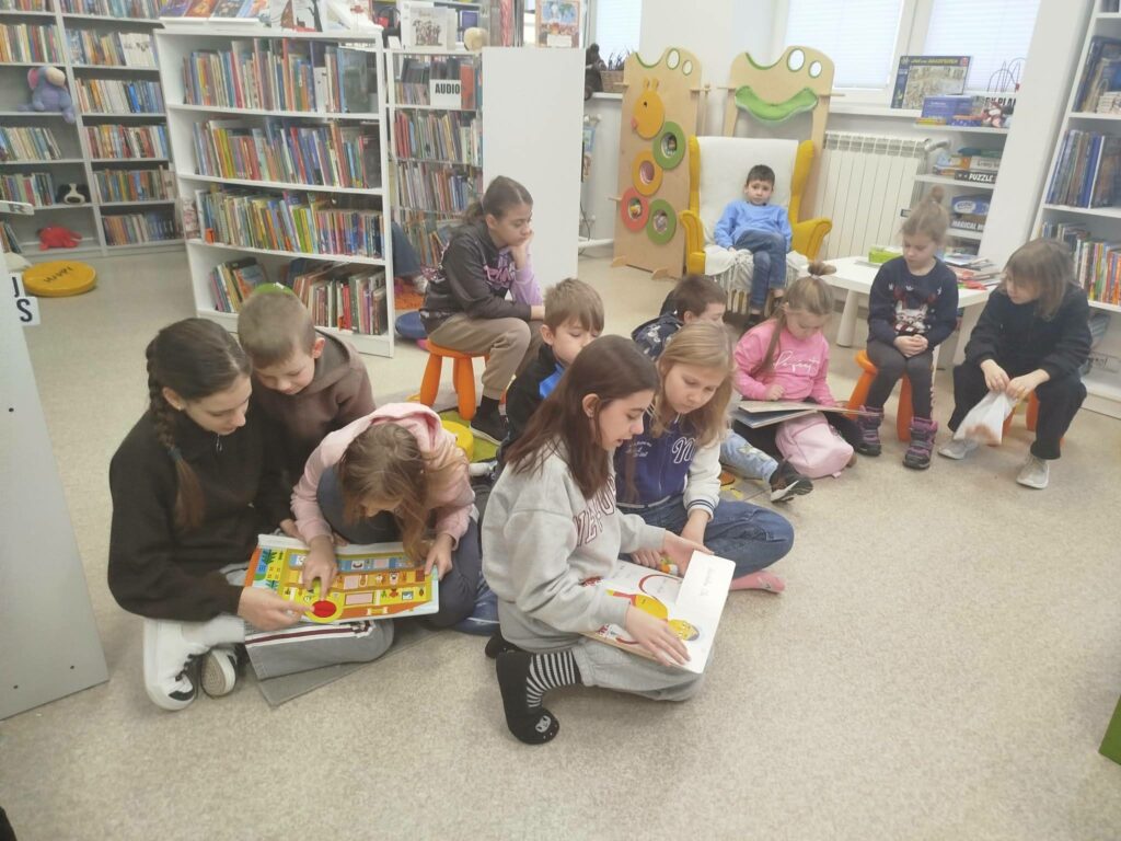 Na ziemi w sali bibliotecznej siedzi grupa dzieci i ogląda ksiązki