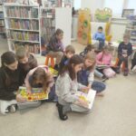 Na ziemi w sali bibliotecznej siedzi grupa dzieci i ogląda ksiązki